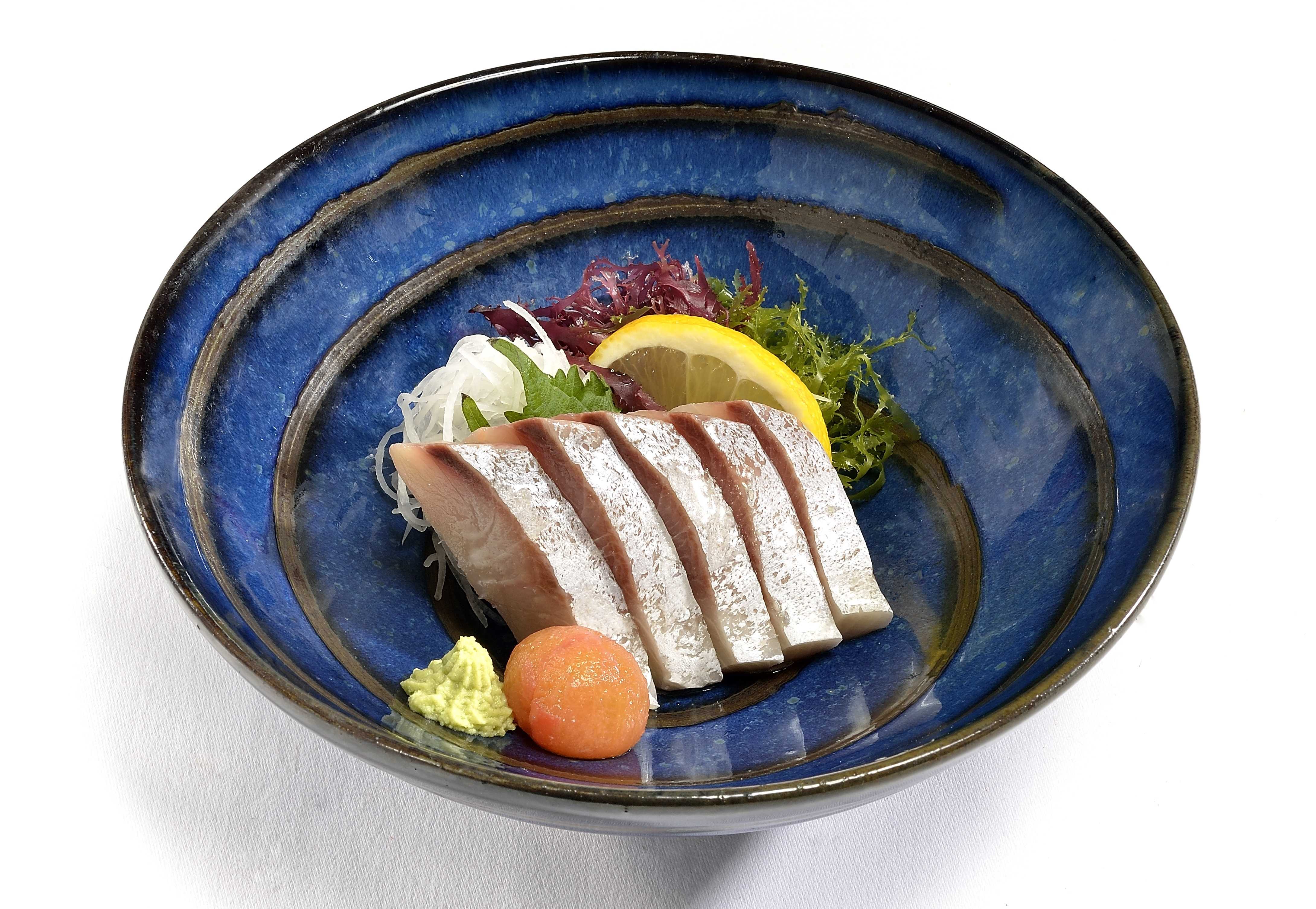 Poisson cru tranché finement avec garniture de citron, algues, wasabi et gingembre râpé dans un bol en céramique bleu profond, ambiance sereine